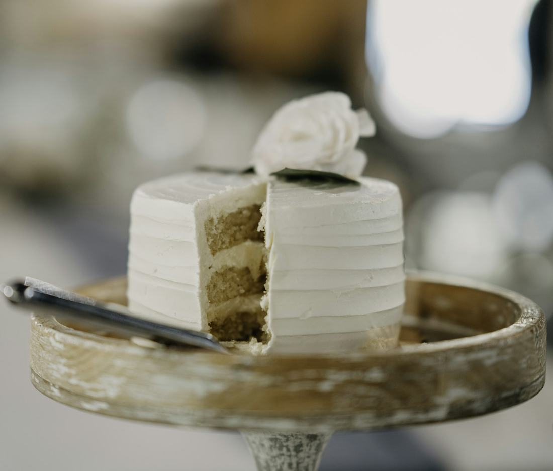 A small white buttercream layer cake with one slice cut away and a single white flower on top, on a weathered wooden cake stand, soft cream and wood tones in diffused light.