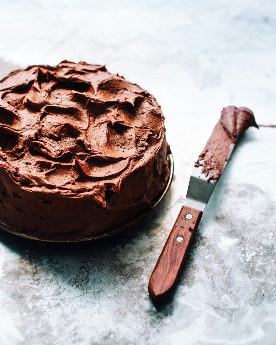 A round cake covered in loose chocolate buttercream swoops beside a wooden handled offset spatula on pale grey blue marble, in soft daylight.