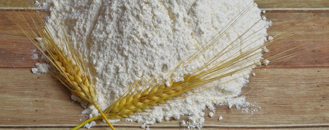 A heap of white flour with two golden wheat ears on worn tan wooden boards, in soft daylight.
