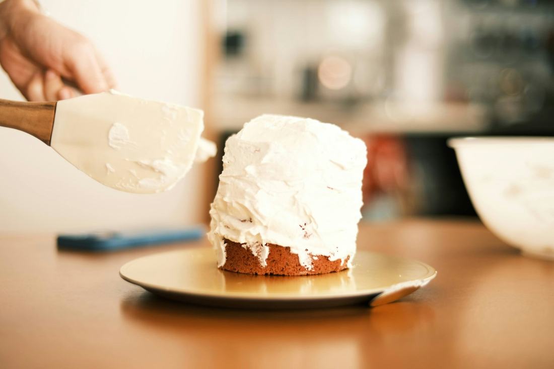 A hand spreading white frosting with a spatula onto a small cake on a gold board, on a warm amber toned kitchen table with a softly blurred background.