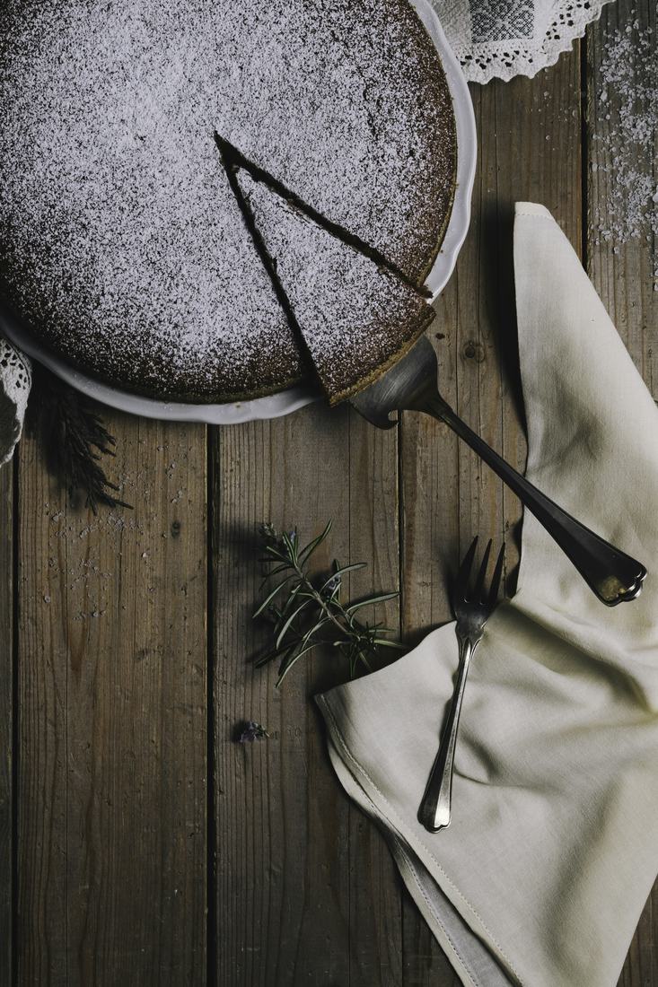 Top down view of a simple sugar dusted cake with a slice cut, a vintage cake server, a fork, a rosemary sprig and a cream linen napkin on dark weathered wood, in moody low light.