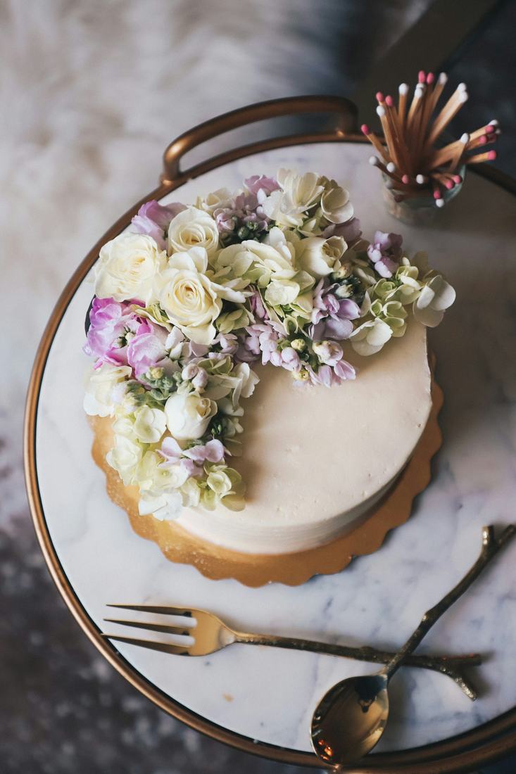 Top down view of a white cake crowned with fresh cream roses, lilac stock and pale green hydrangea on a marble and brass tray with a vintage fork and spoon, in soft natural light.