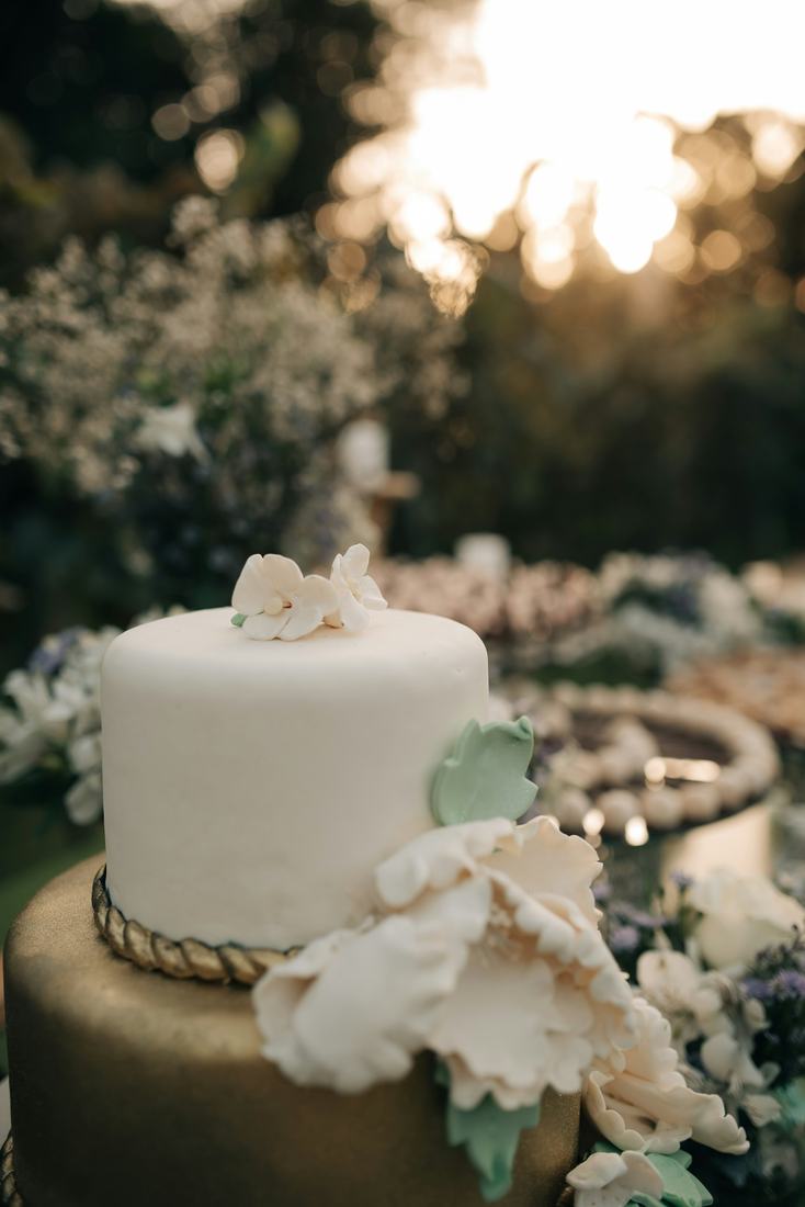 A white cake tier topped with sugar flowers above a bronze lower tier with rope trim and pale sugar peonies, garden blooms behind in low golden sunset light.