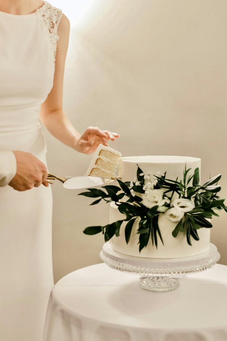 A woman in a white dress, face out of frame, lifting a slice from a white cake dressed with dark green sprigs and white flowers on a glass stand, against cream walls in soft warm light.
