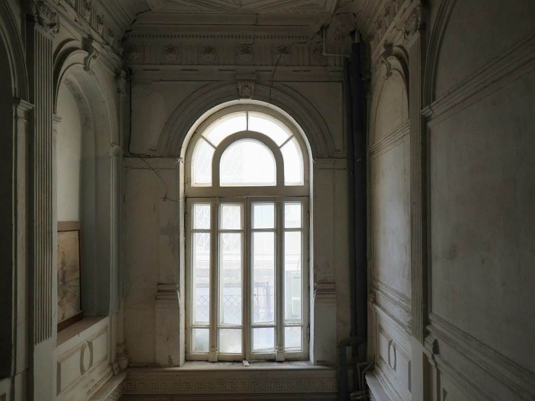 A tall arched window at the end of an old plastered hallway with ornate mouldings, soft daylight falling into muted grey and taupe tones.