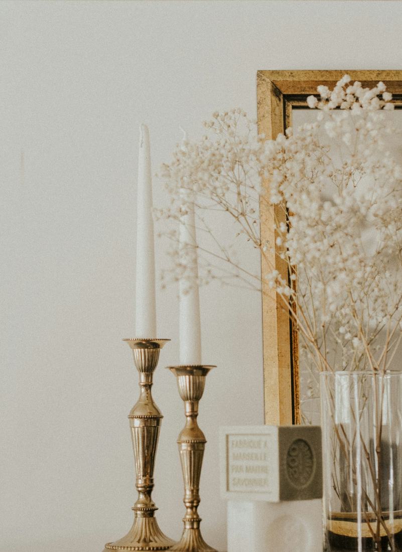 Two antique brass candlesticks with white tapers beside a gilt frame, dried baby's breath and a glass vase against a cream wall, in soft warm light.