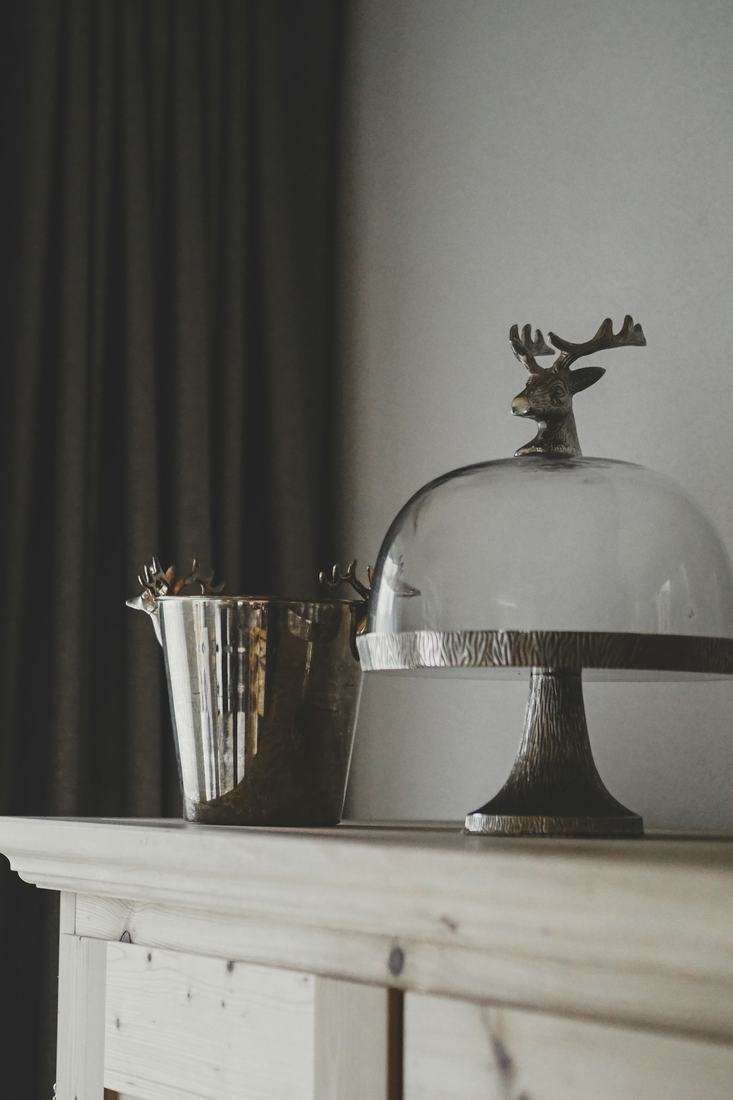 A glass cake dome with a stag head handle on a pewter pedestal beside a silver bucket, on a white wooden cabinet against a grey wall and charcoal curtain, in soft window light.