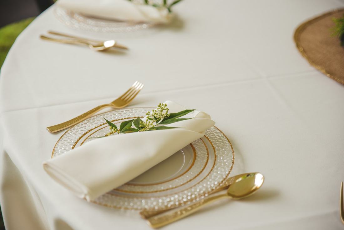 A formal place setting with a folded cream napkin, a green sprig, gold cutlery and a gold ringed glass charger on a white tablecloth, in bright soft light.