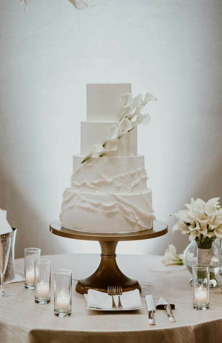 A four tier white cake with soft ruffled icing and cascading white calla lilies on a brass pedestal stand, with votive candles on a cream tablecloth in a bright white room.